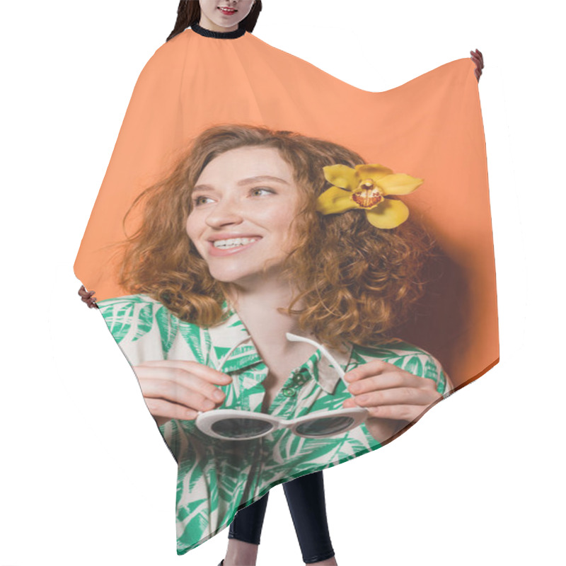 Personality  Smiling Young Redhead Woman With Orchid Flower In Hair Wearing Blouse With Floral Print And Holding Sunglasses While Standing On Orange Background, Summer Casual And Fashion Concept, Youth Culture Hair Cutting Cape