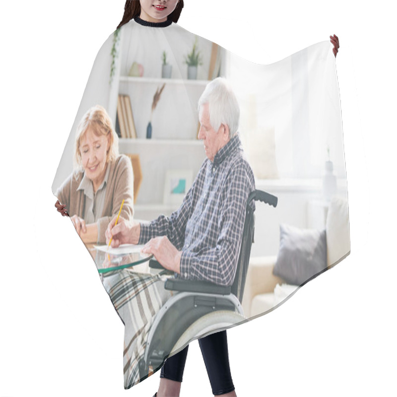 Personality  Aged Disable Man In Casualwear Sitting In Wheelchair By Table And Drawing With Crayons On Paper With His Wife Near By Hair Cutting Cape