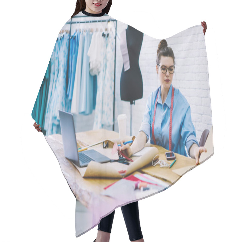 Personality  Young Female Sewing Business Owner Checking Notes In Planner While Working At New Project Sitting At Table With Laptop Drawings And Sewing Supplies In Modern Workshop Hair Cutting Cape