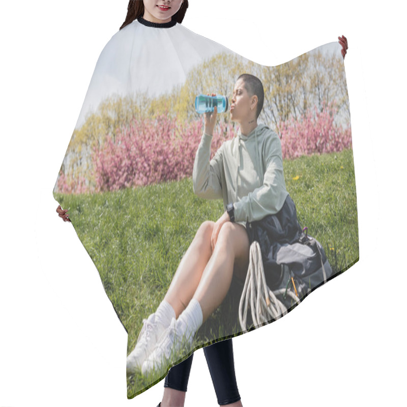 Personality  Young Short Haired And Tattooed Female Backpacker With Smartwatch Drinking Water While Sitting Near Backpack On Grassy Lawn With Nature At Background, Trailblazing Through Scenic Landscape Hair Cutting Cape