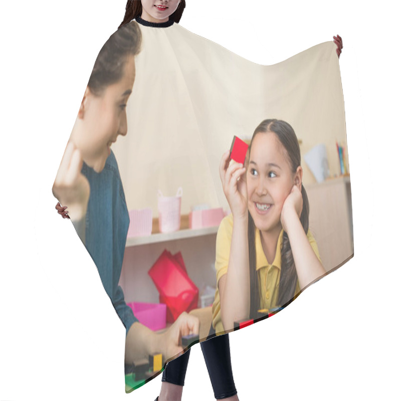 Personality  Cheerful Asian Girl Holding Wooden Cube Near Tattooed Teacher Hair Cutting Cape