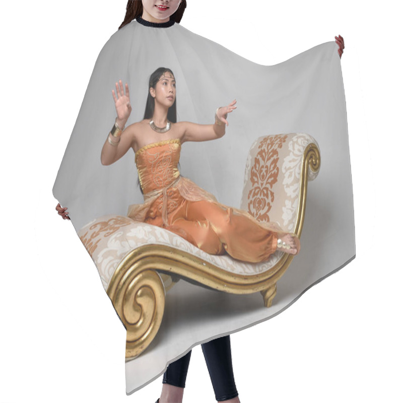 Personality  Full Length Portrait Of Pretty Young Asian Woman Wearing Golden Arabian Robes Like A Genie, Seated Pose On Lounge, Isolated On Studio Background. Hair Cutting Cape