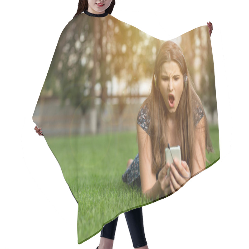 Personality  Anxious Young Girl Looking At Phone Seeing Bad News Or Photos There With Frustrated Disgusting Emotion On Her Face Outdoors Lying Down On Green Lawn Headphones On Head. Multicultural Model, Mixed Race Hair Cutting Cape