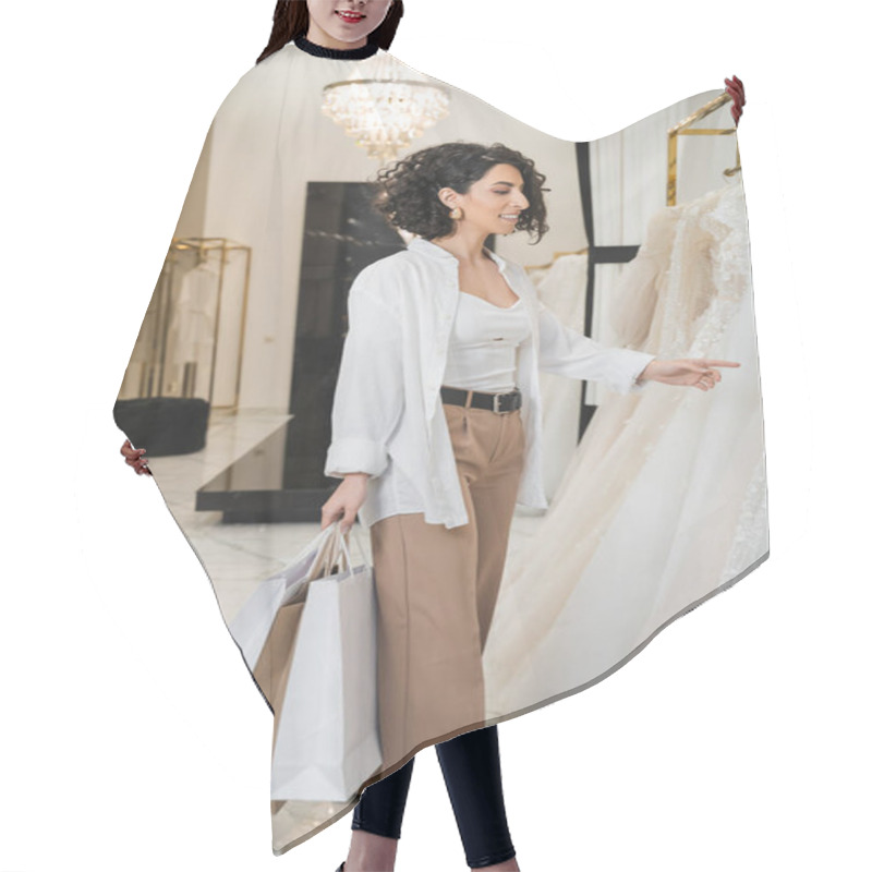 Personality  Stylish And Happy Middle Eastern Woman With Brunette And Wavy Hair Standing In Beige Pants With White Shirt And Holding Shopping Bags While Choosing Wedding Dress In Bridal Salon  Hair Cutting Cape