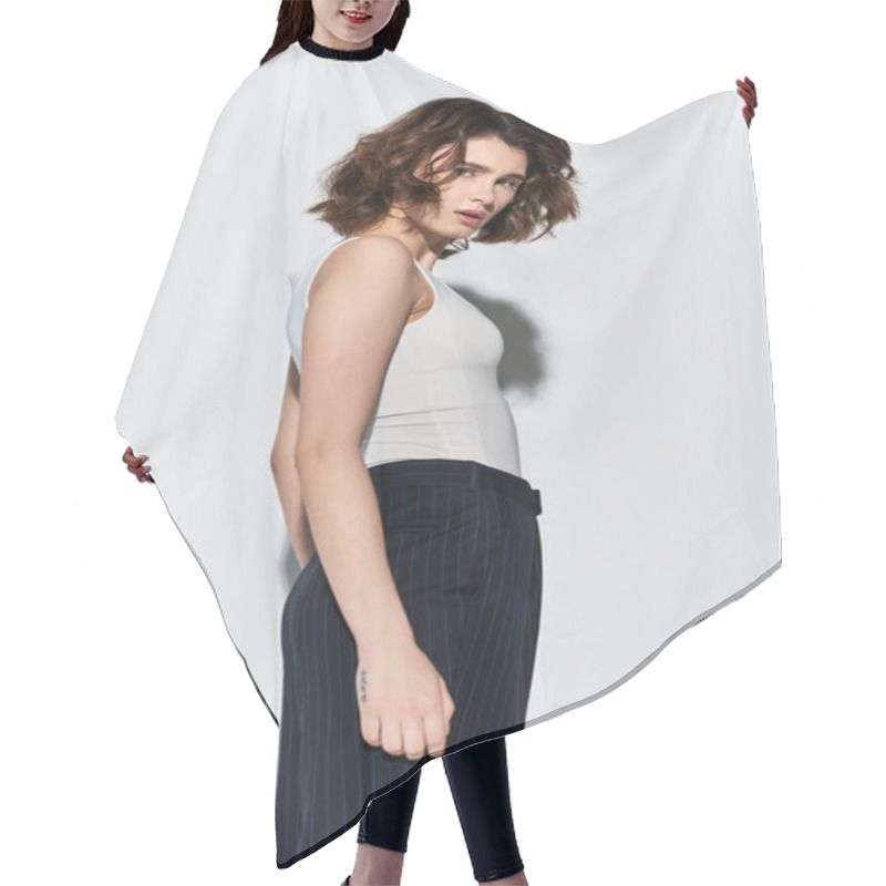 Personality  A Young, Pretty Woman Poses Elegantly In A White Tank Top And Black Pants Against A Grey Studio Backdrop. Hair Cutting Cape