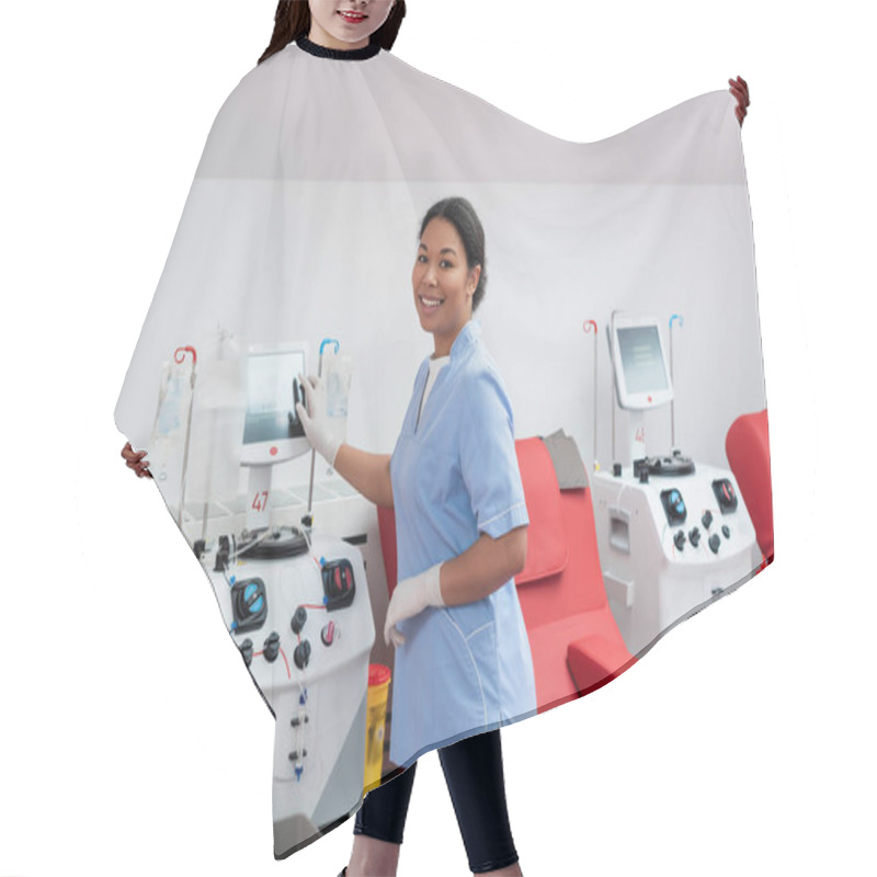 Personality  Young Multiracial Nurse In Uniform And Latex Gloves Operating Transfusion Machine And Smiling At Camera Near Medical Chairs In Blood Donation Center Hair Cutting Cape