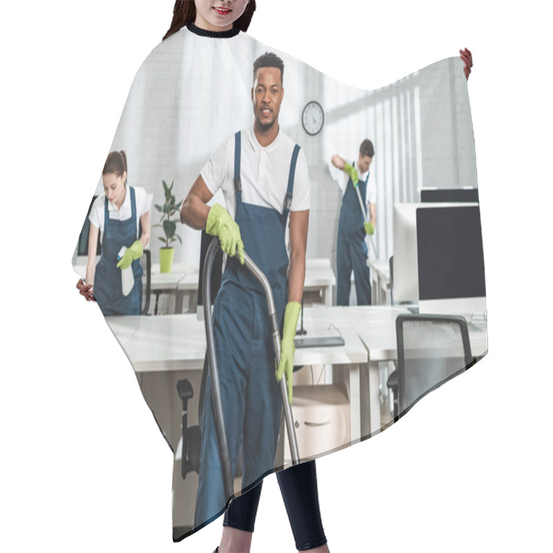 Personality  Handsome African American Cleaner Looking At Camera While Standing With Vacuum Cleaner Near Colleagues Hair Cutting Cape