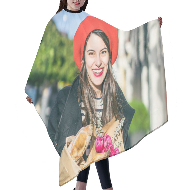 Personality  Young Beautiful Girl Frenchwoman Brunette In A Red Beret And A Black Coat Goes Along The Street Of The European City With A Bouquet Of Flowers And French Baguettes. A Happy Smile And A Charming Look, The French Style Of Paris Hair Cutting Cape