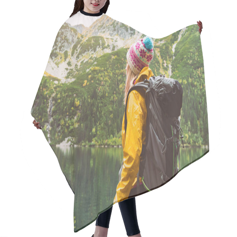 Personality  Young Blond Hair Woman With Big Camping Rucksack Wearing Yellow Raincoat Jacket, Purple And Blue Cap Looking On Mountains And Lake. Copy Space, Rear View Background. Hair Cutting Cape