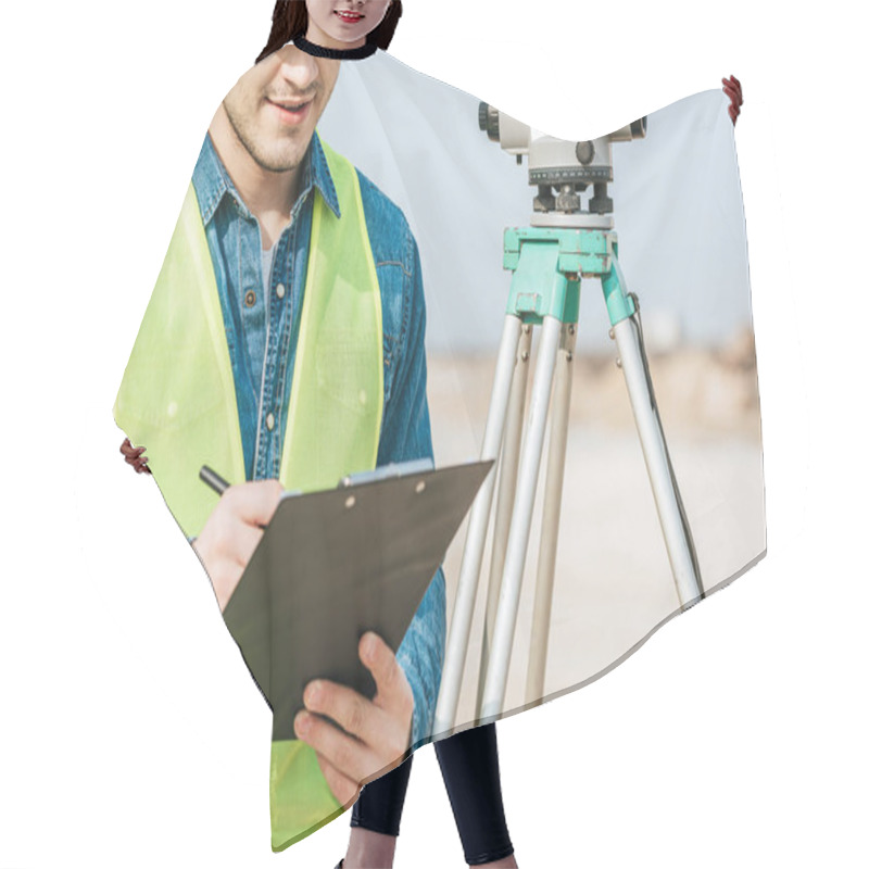 Personality  Cropped Shot Of Smiling Surveyor Writing On Clipboard Beside Digital Level  Hair Cutting Cape