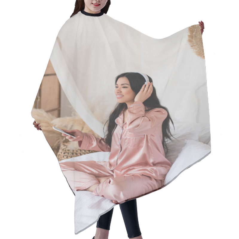 Personality  Young Asian Woman In Pink Silk Pajamas Sitting On Bed With Cellphone And Touching Headphones With Hand In Bedroom Hair Cutting Cape