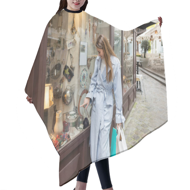 Personality  Cheerful Woman In Trench Coat Standing With Shopping Bags And Pointing At Showcase Of Antique Shop In Vienna  Hair Cutting Cape