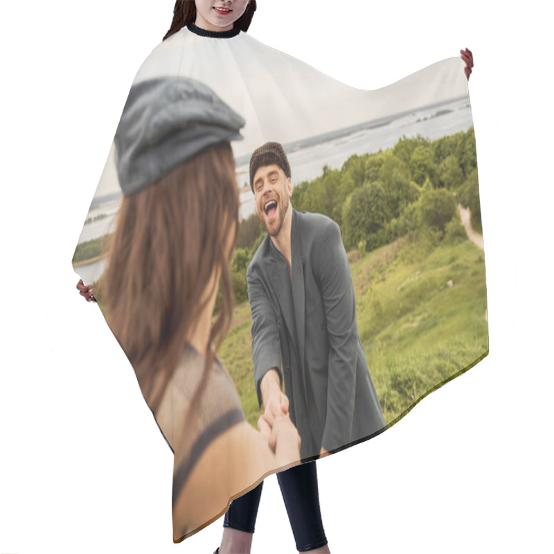 Personality  Cheerful And Trendy Bearded Man In Jacket And Newsboy Cap Holding Hand Of Blurred Girlfriend While Standing With Scenic Landscape And Sky At Background, Fashion-forwards In Countryside Hair Cutting Cape