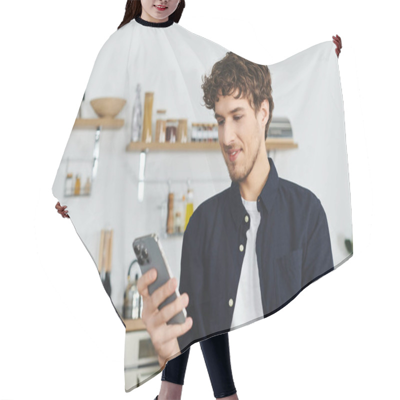 Personality  Curly-haired Man In A Stylish Shirt Smiles As He Scrolls Through His Smartphone At Home. Hair Cutting Cape