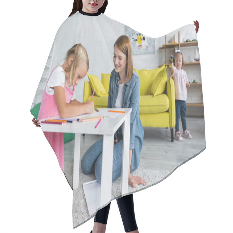 Personality  Happy Kindergarten Teacher Looking At Girl Drawing While Toddler Kid With Down Syndrome Standing On Blurred Background Hair Cutting Cape