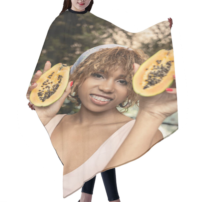 Personality  Cheerful Young African American Woman With Braces Wearing Summer Dress And Headscarf While Holding Cut Papaya And Looking At Camera In Orangery, Fashion-forward Lady Inspired By Tropical Plants Hair Cutting Cape