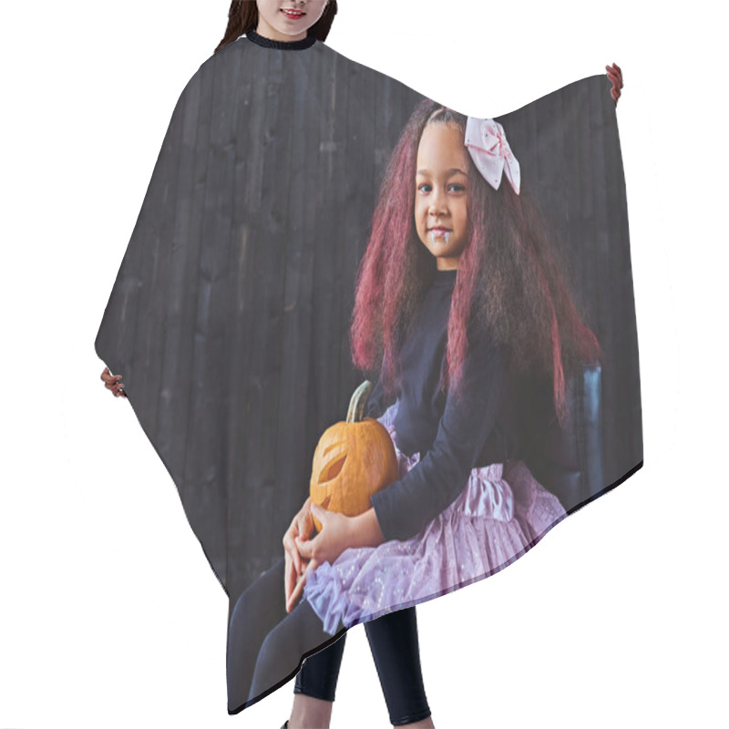 Personality  Dark-skinned Little Girl Dressed In Trendy Scary Dress Holds Pumpkin While Sitting On Chair In Dark House. Halloween Concept. Hair Cutting Cape