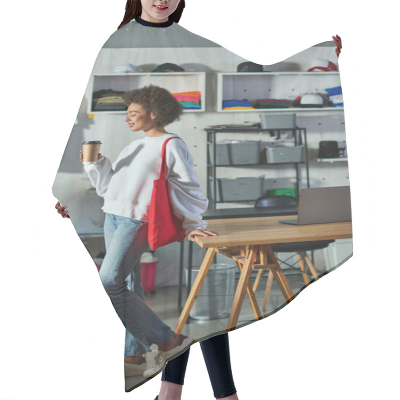 Personality  Side View Of Young And Positive African American Craftswoman With Shoulder Bag Holding Coffee To Go And Standing Near Laptop On Table In Print Studio, Enthusiastic Business Owner Working In Workshop Hair Cutting Cape