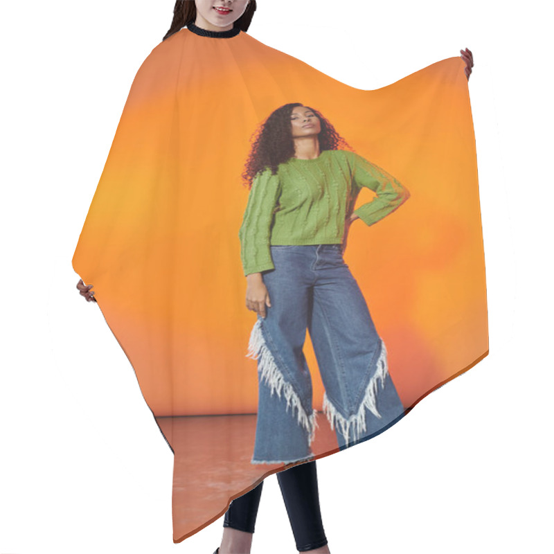 Personality  Woman In A Green Sweater And Fringed Jeans Strikes A Pose Against An Orange Backdrop. Hair Cutting Cape