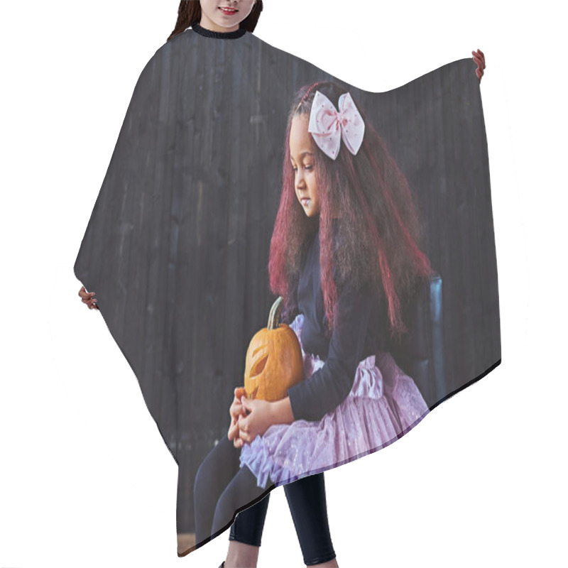 Personality  Dark-skinned Little Girl Dressed In Trendy Scary Dress Holds Pumpkin While Sitting On Chair In Dark House. Halloween Concept. Hair Cutting Cape