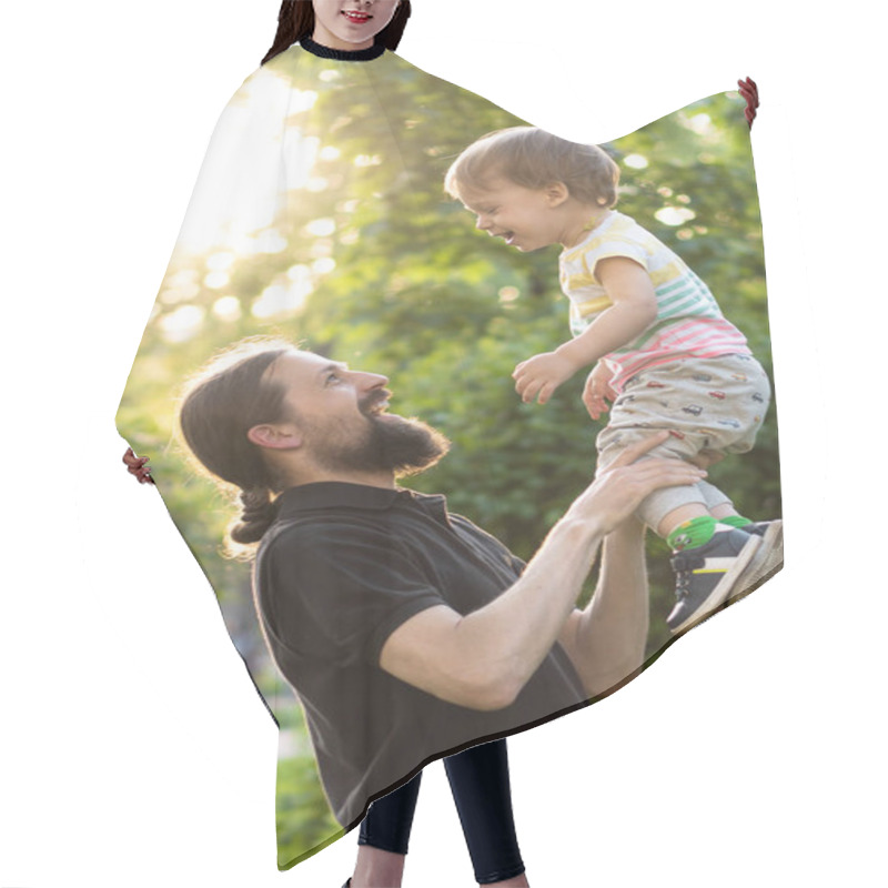 Personality  Fatherhood, Parenthood, Childhood, Caring, Summer And Leisure Concept - Young Dad With Beard And Long Hair In Black T-shirt Holds In His Arms Little Son In The Backlight Of The Sunset In The Park. Hair Cutting Cape