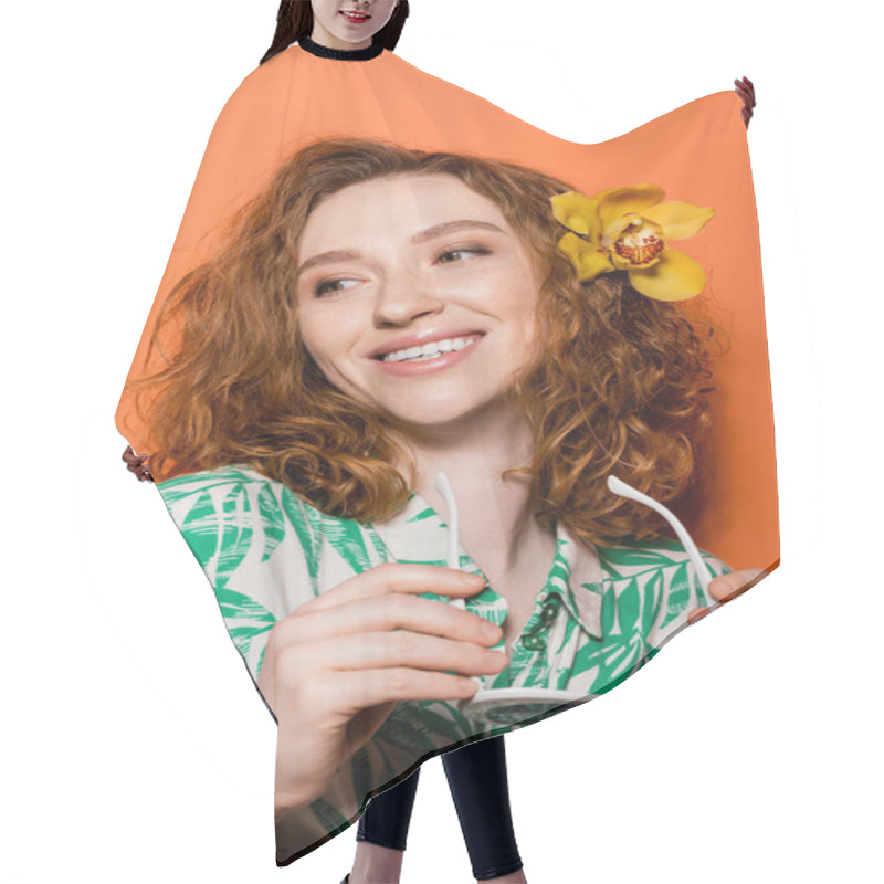 Personality  Positive Young Redhead Woman With Orchid Flower In Hair Wearing Blouse With Floral Pattern And Holding Sunglasses On Orange Background, Summer Casual And Trendy Concept, Youth Culture Hair Cutting Cape