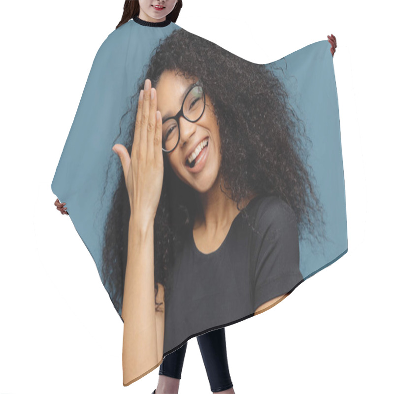 Personality  Happy Afro American Woman Touches Forehead, Tilts Head, Smiles Happily At Camera, Has Fun Indoor, Wears Optical Spectacles And Casual Black T Shirt, Isolated Over Blue Background. Emotions Concept Hair Cutting Cape