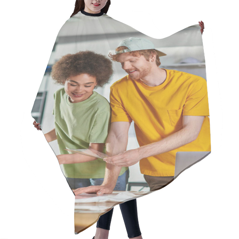 Personality  Smiling Craftsman Holding Printing Layer Near African American Colleague, Clothes And Laptop On Table While Working In Blurred Print Studio At Background, Start-up Innovation Concept Hair Cutting Cape