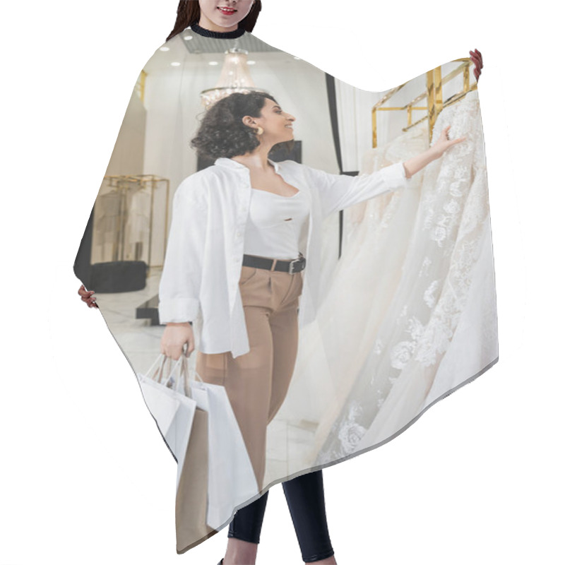 Personality  Stylish And Joyful Middle Eastern Woman With Brunette And Wavy Hair Standing In Beige Pants With White Shirt And Holding Shopping Bags While Choosing Wedding Dress In Bridal Boutique, Chic  Hair Cutting Cape