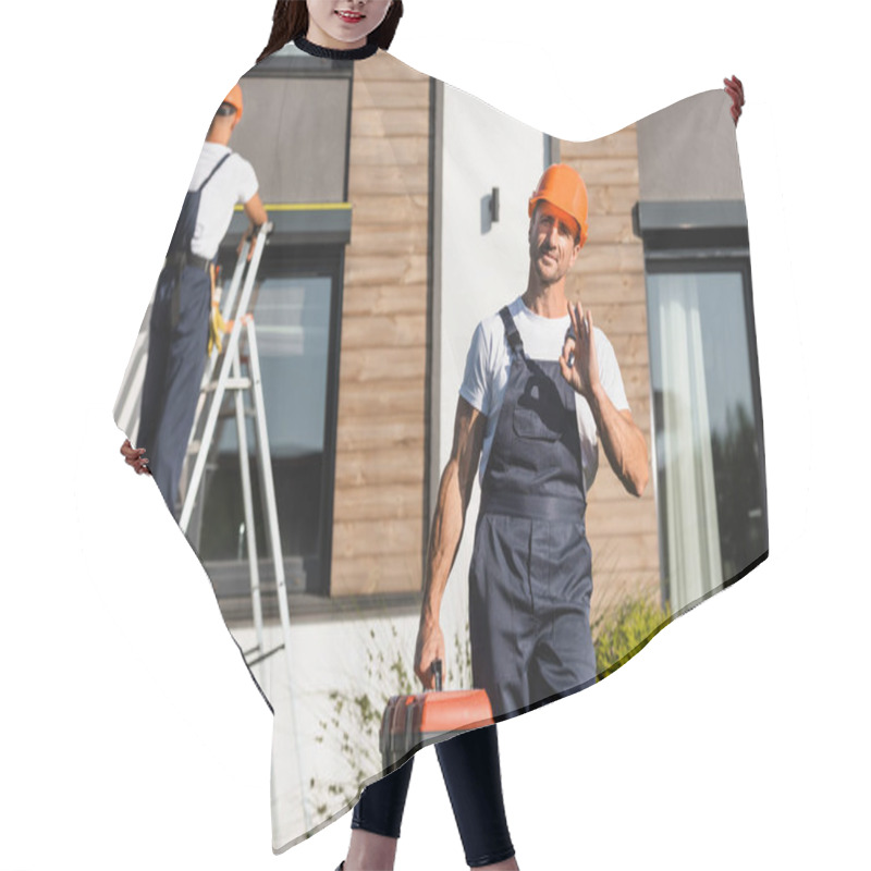 Personality  Selective Focus Of Builder In Uniform With Toolbox Showing Ok Gesture While Colleague Working Near Building  Hair Cutting Cape