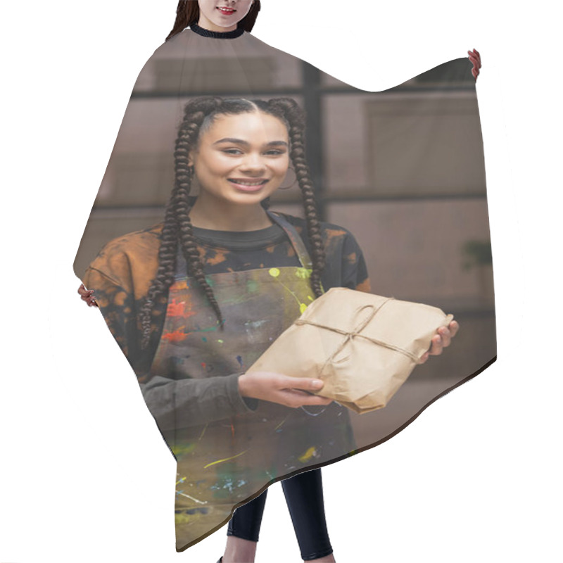 Personality  Positive African American Designer In Apron Holding Package And Looking At Camera In Workshop  Hair Cutting Cape