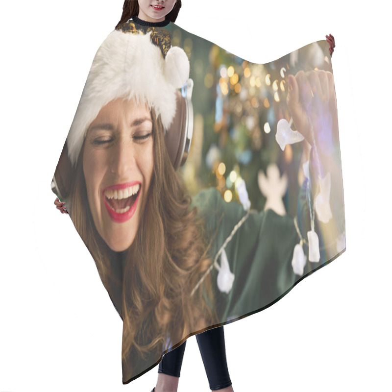 Personality  Christmas Time. Smiling Trendy Female With Santa Hat In Green Dress Listening To The Music With Headphones And Dancing Near Christmas Tree In The Modern House. Hair Cutting Cape