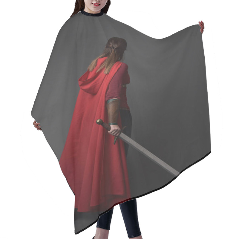 Personality  Full Length Portrait Of Brunette Girl Wearing Red Medieval Costume And Cloak. Standing Pose  With Back To The Camera, Holding A Sword On Grey Studio Background. Hair Cutting Cape