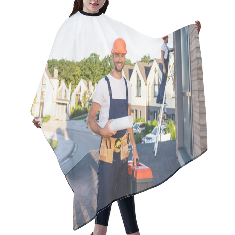 Personality  Selective Focus Of Builder With Toolbox And Blueprint Looking At Camera While Colleague Working On Roof  Hair Cutting Cape