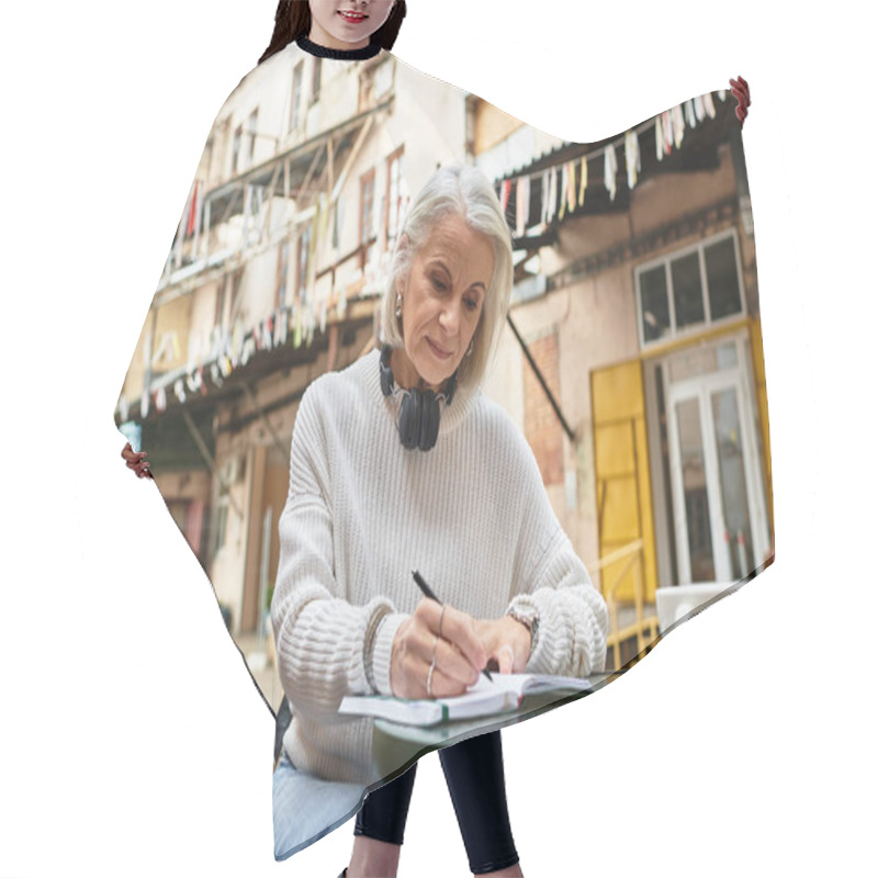 Personality  The Beautiful Gray Haired Woman Sits At A Table, Writing Attentively In A Lively Courtyard. Hair Cutting Cape