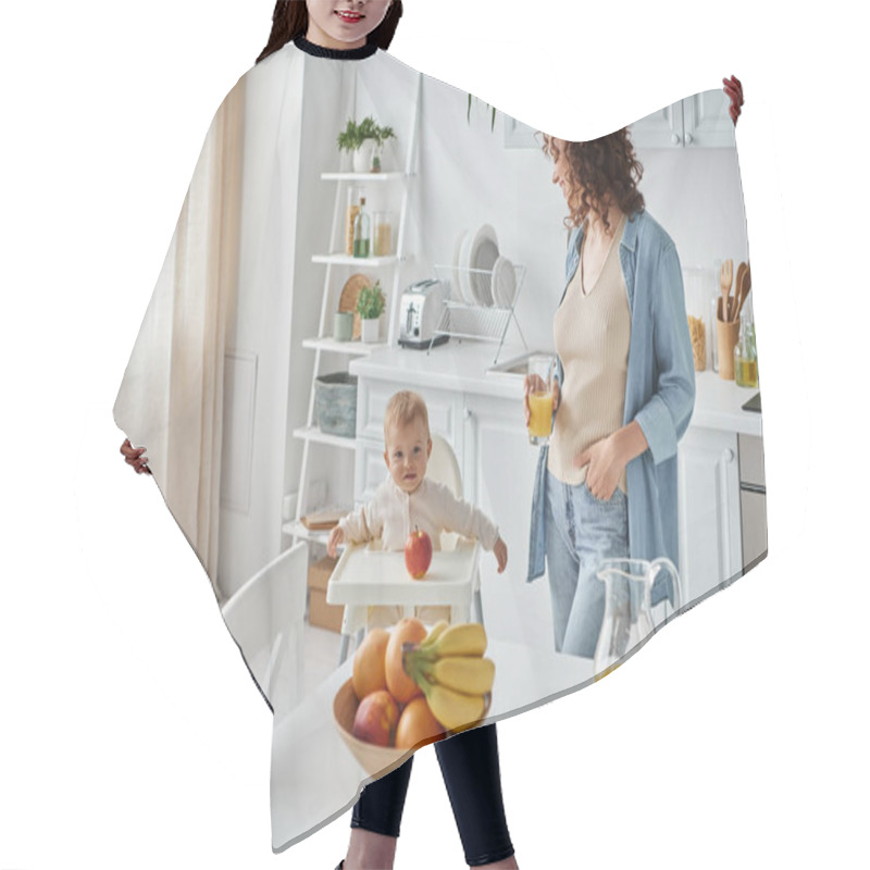 Personality  Curly Woman With Glass Of Orange Juice Looking At Child In Baby Chair In Kitchen With Ripe Fruits Hair Cutting Cape