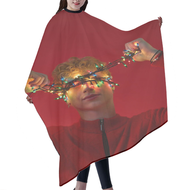 Personality  A Young Man With Curly Hair Playfully Poses Against A Red Backdrop, Wrapped In Colorful Christmas Lights. Hair Cutting Cape