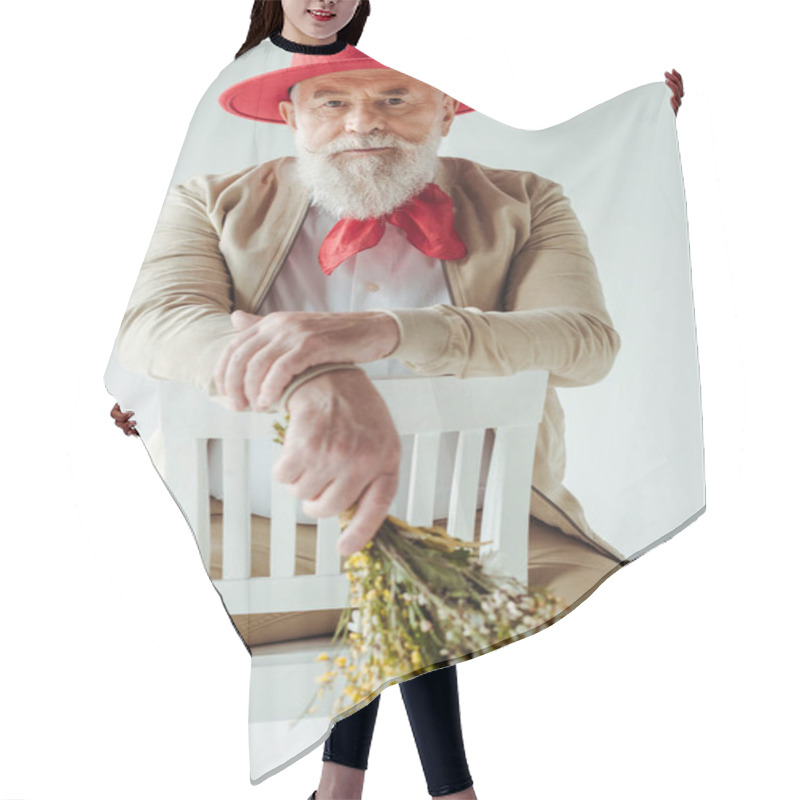 Personality  Selective Focus Of Stylish Senior Man In Red Hat Holding Wildflowers And Looking At Camera On Chair Isolated On White Hair Cutting Cape