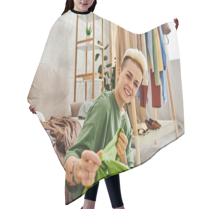 Personality  Young Excited Woman Sorting Second-hand Clothes And Holding Green Garment Near Couch And Rack At Home, Trendy Hairstyle, Tattoo, Looking At Camera, Sustainable Living And Mindful Consumerism Concept Hair Cutting Cape