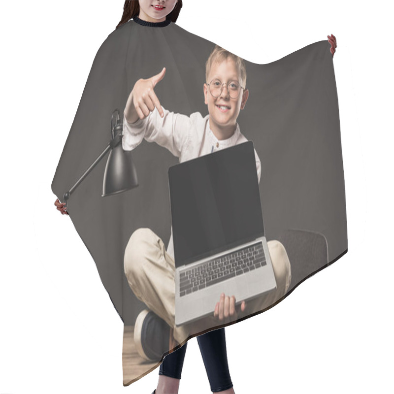 Personality  Smiling Little Boy In Eyeglasses Sitting On Table And Pointing On Laptop With Blank Screen On Grey Background  Hair Cutting Cape