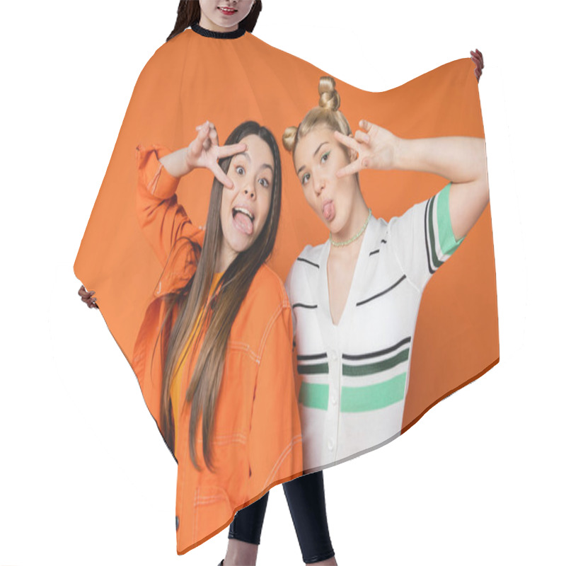 Personality  Portrait Of Trendy Teenage Girlfriends With Bright Makeup Sticking Out Tongues And Showing Peace Gesture At Camera While Standing Isolated On Orange, Fashionable Girls With Sense Of Style Hair Cutting Cape