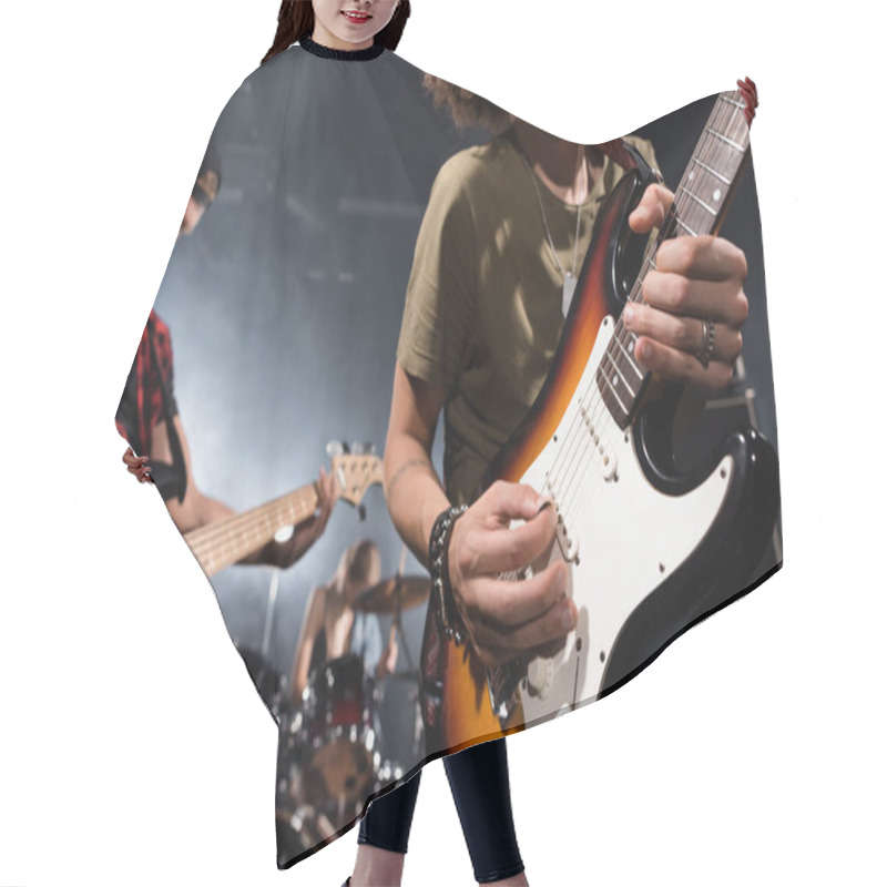 Personality  KYIV, UKRAINE - AUGUST 25, 2020: Curly Musician Playing Electric Guitar With Guitar Pick With Blurred Musicians On Background Hair Cutting Cape