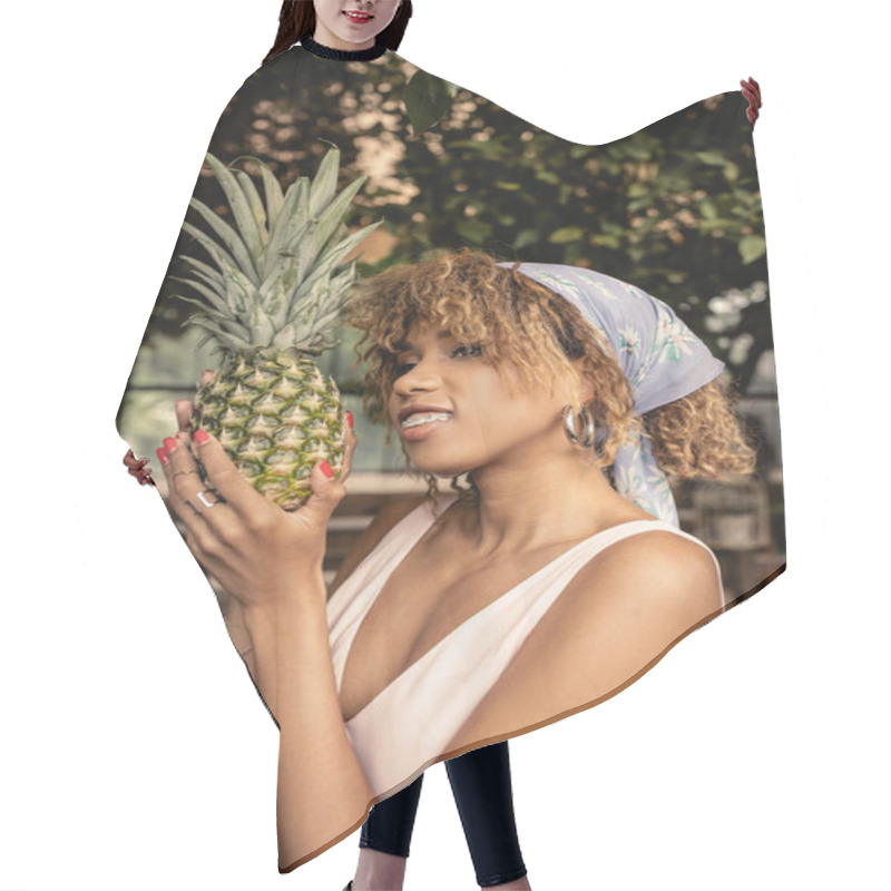 Personality  Portrait Of Smiling And Stylish African American Woman With Braces In Headscarf Holding Fresh Pineapple And Looking Away While Standing In Blurred Greenhouse, Fashion-forward Lady In Tropical Oasis Hair Cutting Cape