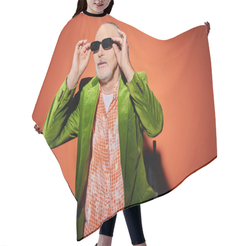 Personality  Positive And Fashionable Aging Concept, Senior Male Model In Green Velour Blazer And Trendy Shirt Looking Away And Adjusting Dark Sunglasses While Sitting On Chair On Red And Orange Background Hair Cutting Cape