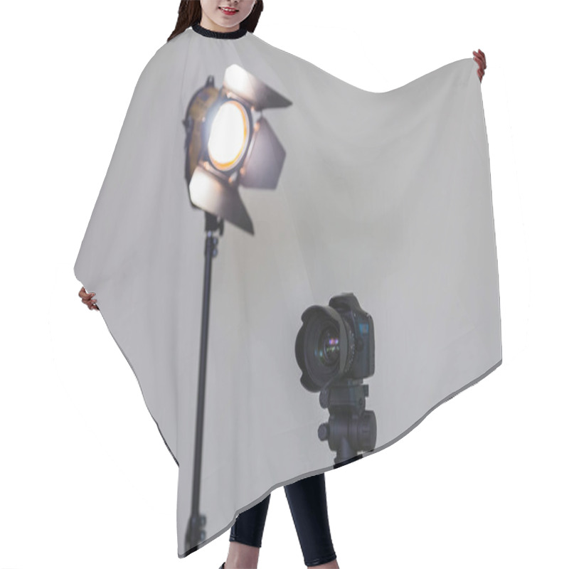 Personality  Digital SLR Camera And A Spotlight With A Fresnel Lens On A Gray Background. Shooting In The Interior. Equipment For The Production Of Films Hair Cutting Cape