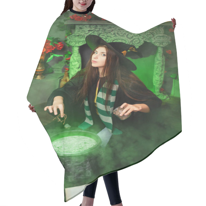 Personality  Attractive Girl In The Image Of A Witch Dressed In A Black Hat And Mantle Throws An Ingredient Into A Cauldron With An Upcoming Potion, Sitting In A Magical, Green Smoke. Halloween Witch Concept. Hair Cutting Cape