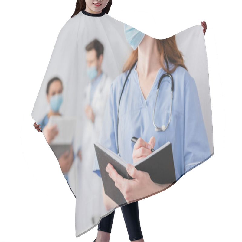 Personality  Redhead Nurse In Medical Mask Writing In Notebook With Blurred Multiethnic Colleagues Working On Background Hair Cutting Cape