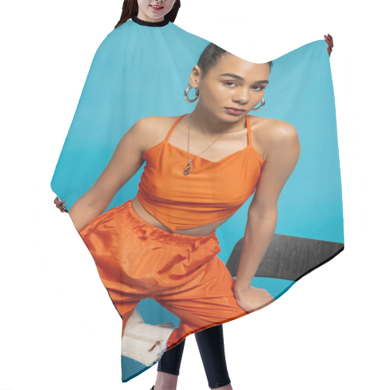 Personality  Beautiful Fashion Model In Orange Outfit Posing On Tall Chair Looking At Camera, Blue Background Hair Cutting Cape