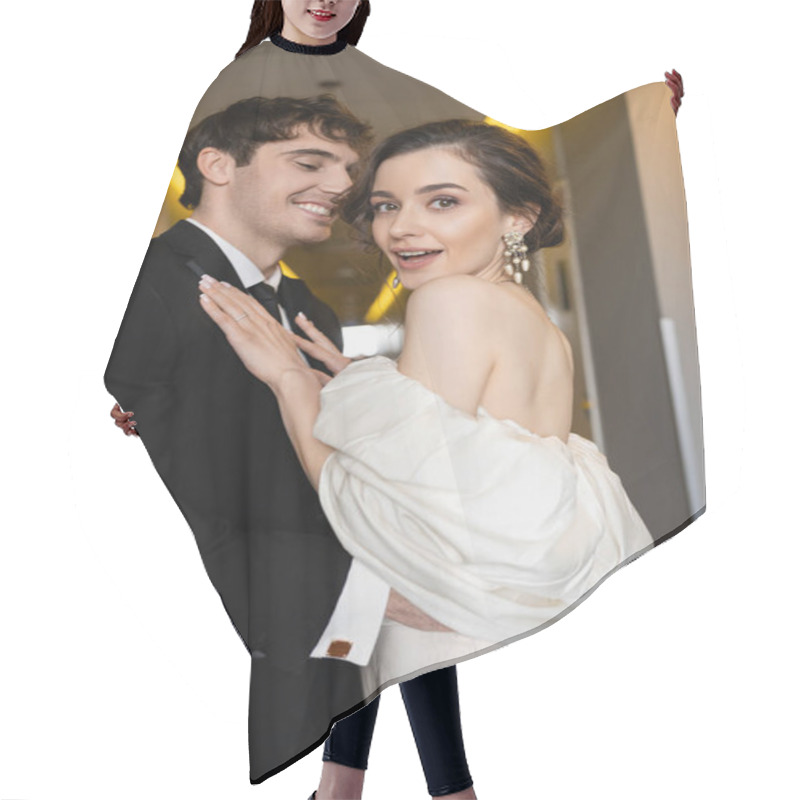 Personality  Happy Groom In Black Suit Hugging Excited Bride In White Wedding Dress And Pearl Earrings While Smiling Together In Corridor Of Modern Hotel, Couple On Honeymoon   Hair Cutting Cape