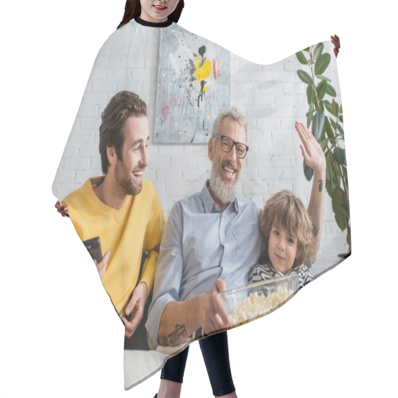 Personality  Smiling Man With Popcorn Waving Hand Near Son With Remote Controller And Grandson On Couch  Hair Cutting Cape